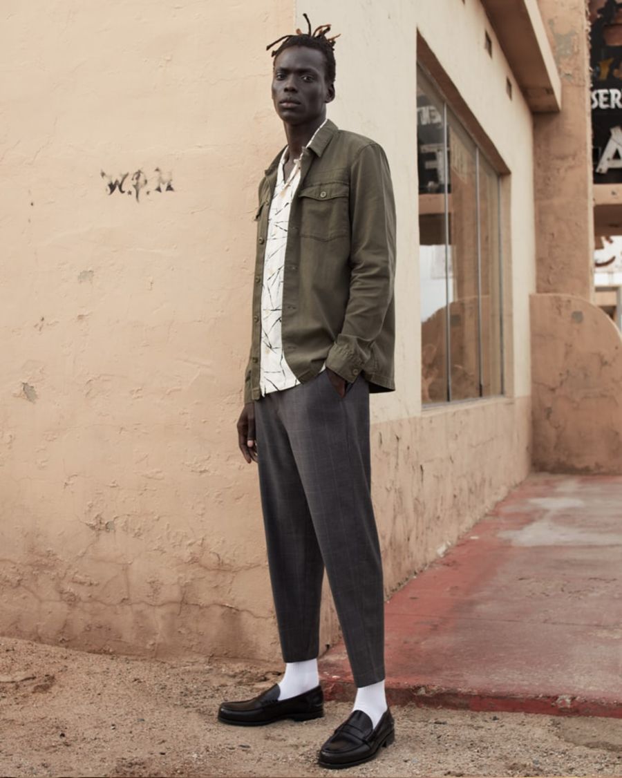 Foto von einem Mann, der in einer Straße steht und eine khakifarbene Jacke über einem bedruckten Hemd und einer grauen Hose trägt.