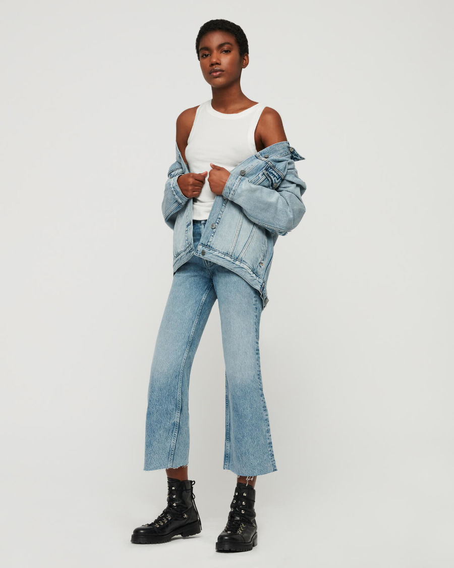 Image of woman standing in front of a white wall wearing a pair of light high waisted jeans with a denim jacket worn on top of a white tank.