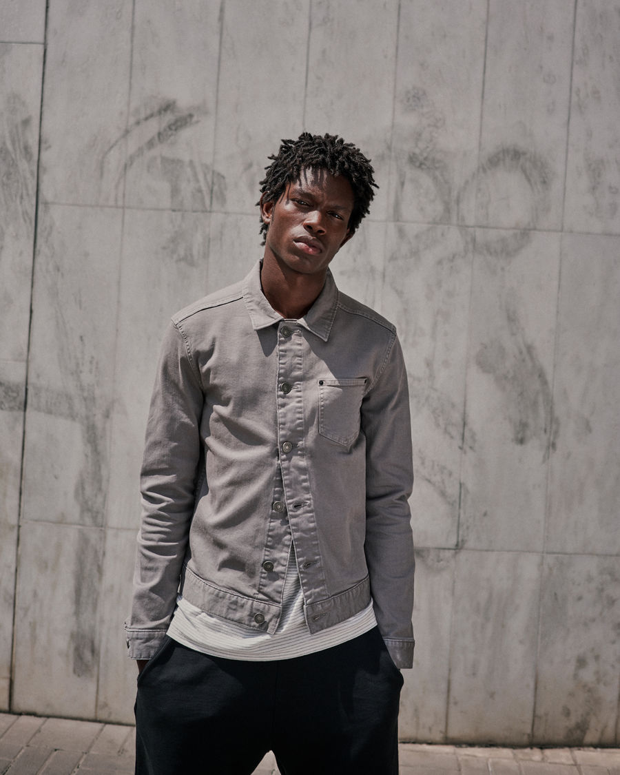 Image of a man wearing a grey denim jacket on top of a stripy t-shirt worn with dark sweatpants.