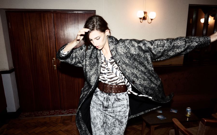 Image of a woman in a corridor wearing a grey textured pencil skirt with a brown leather belt, a zebra printed shirt and a black and white coat.