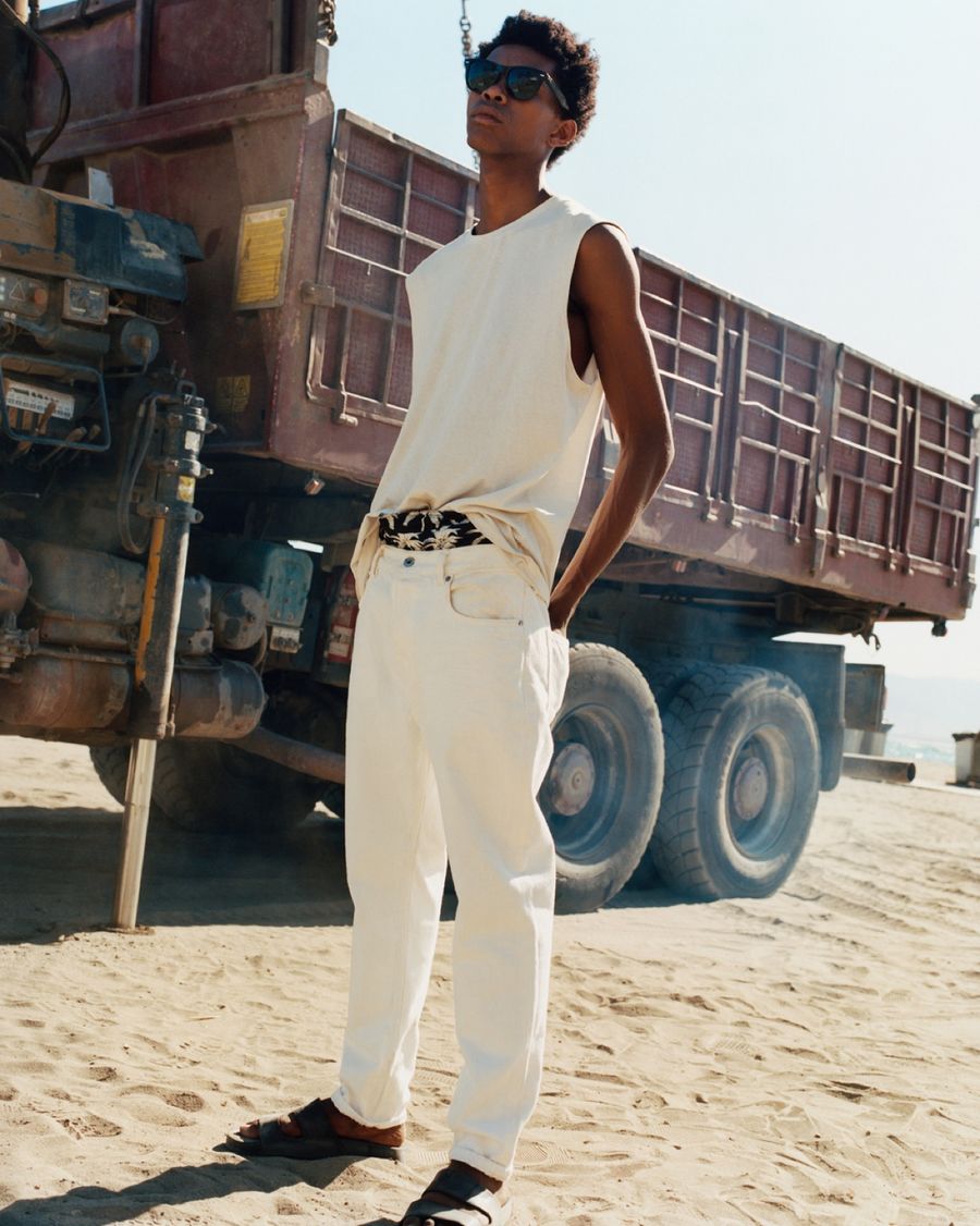 Image of a man standing in front of a truck wearing a white vest and a palm tree printed pair of shorts under white jeans with black leather slides.