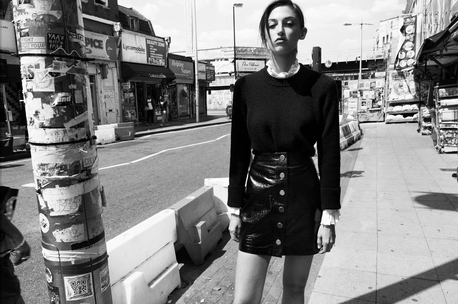 Black and white image of a woman walking in the street wearing a mini leather skirt and black jumper over a white shirt.