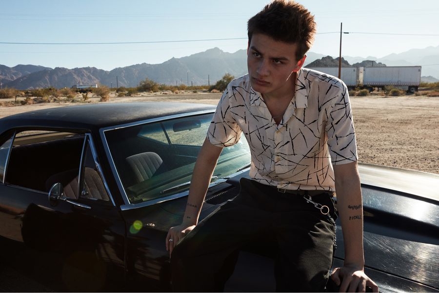 Portrait of a man sitting on a black sports car wearing a black and white printed shirt with black tailored trousers.