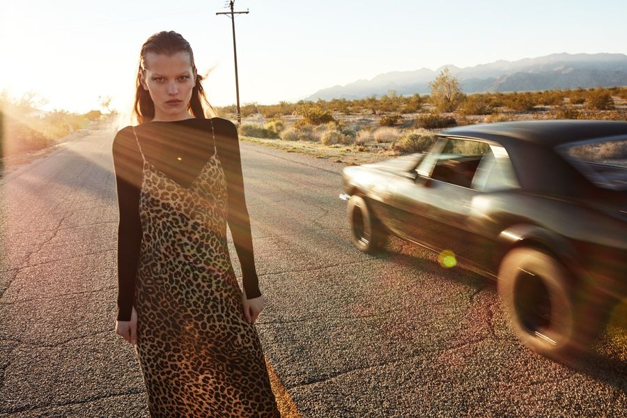Image of a woman standing on the road near a black sport car and wearing a thin strap leopard print dress over a thin long sleeve jumper.