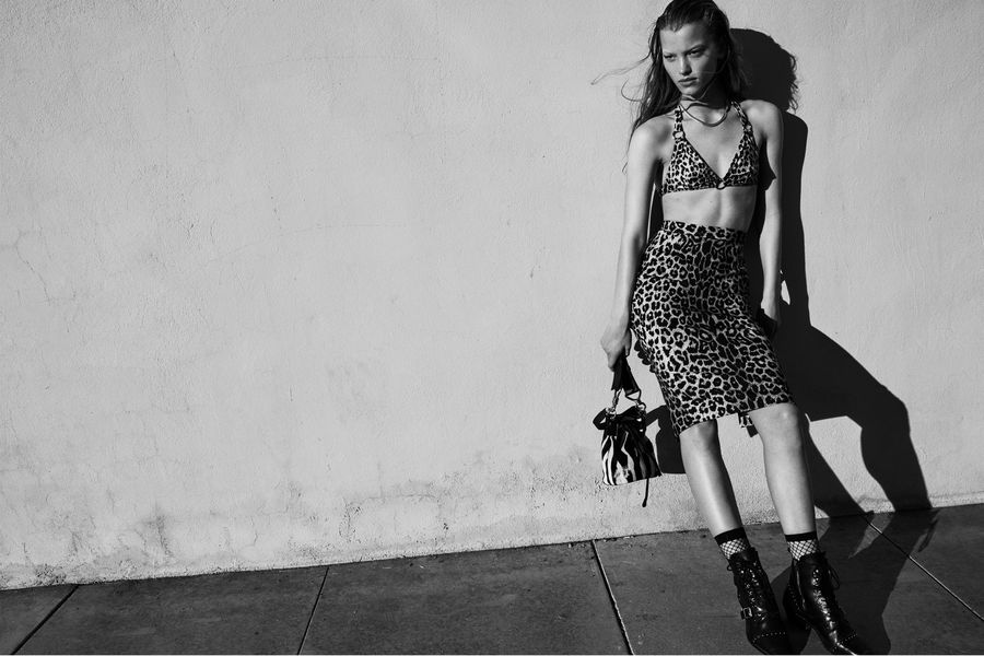 Black and white image of a woman wearing a leopard print bikini top and matching pencil skirt with black boots and a zebra printed bucket bag.