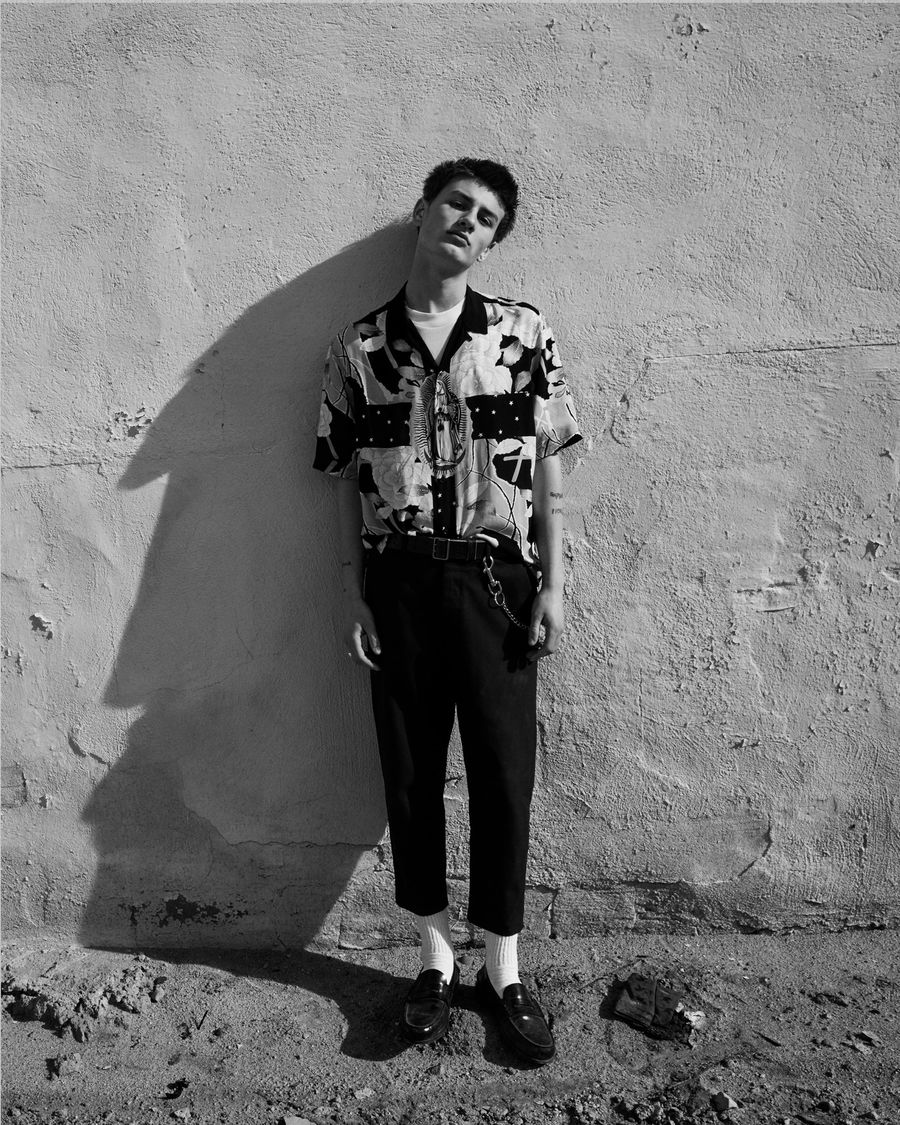 Black and white portrait of a man standing against a wall and wearing a printed shirt over a white t-shirt with black tailored trousers.