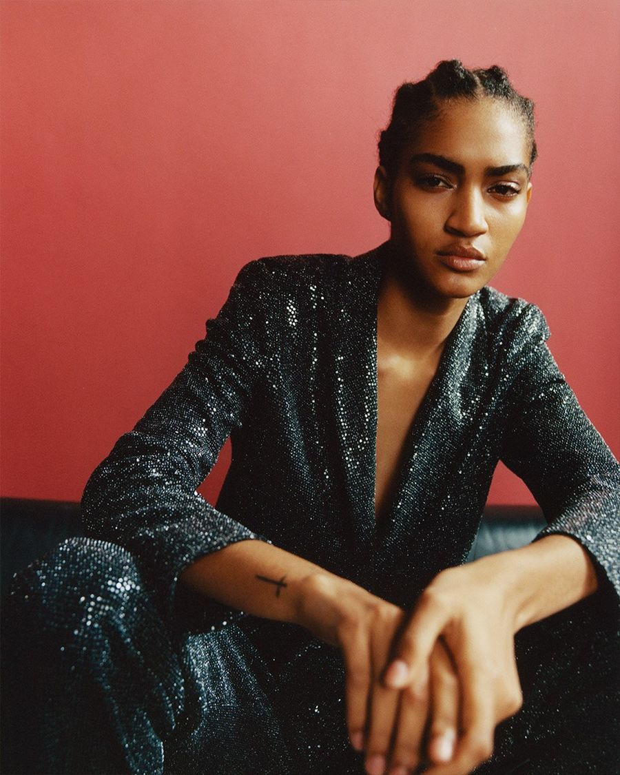 Immagine di una donna seduta su una superficie in pelle di fronte ad un muro rosso. Indossa un completo con paillettes all-over.