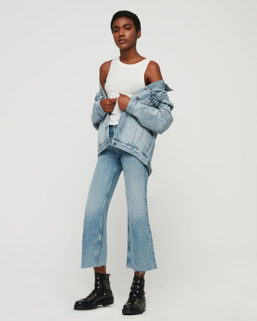 Immagine di una donna ferma di fronte a un muro bianco. Indossa jeans a vita alta con una giacca e una canotta bianca.