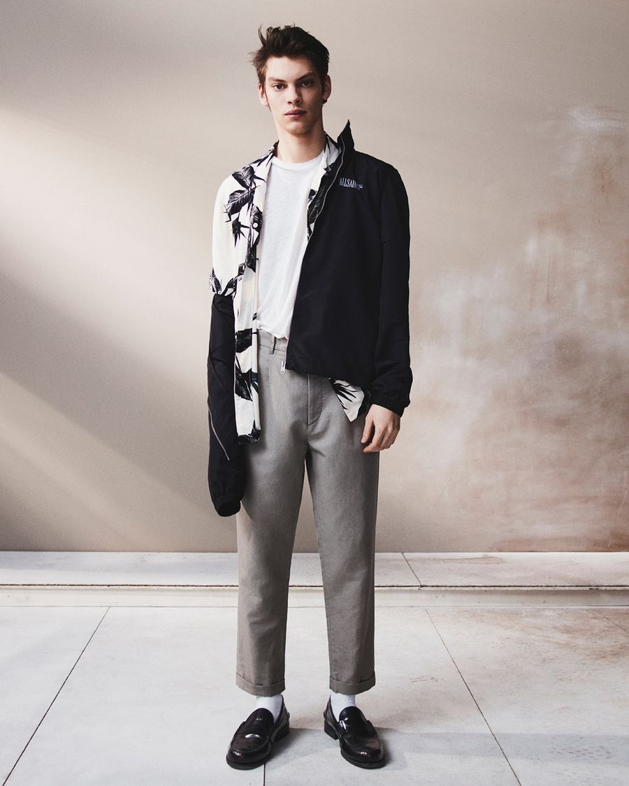 Imagen en blanco y negro de un hombre con un abrigo largo gris, pantalones negros y mocasines negros.