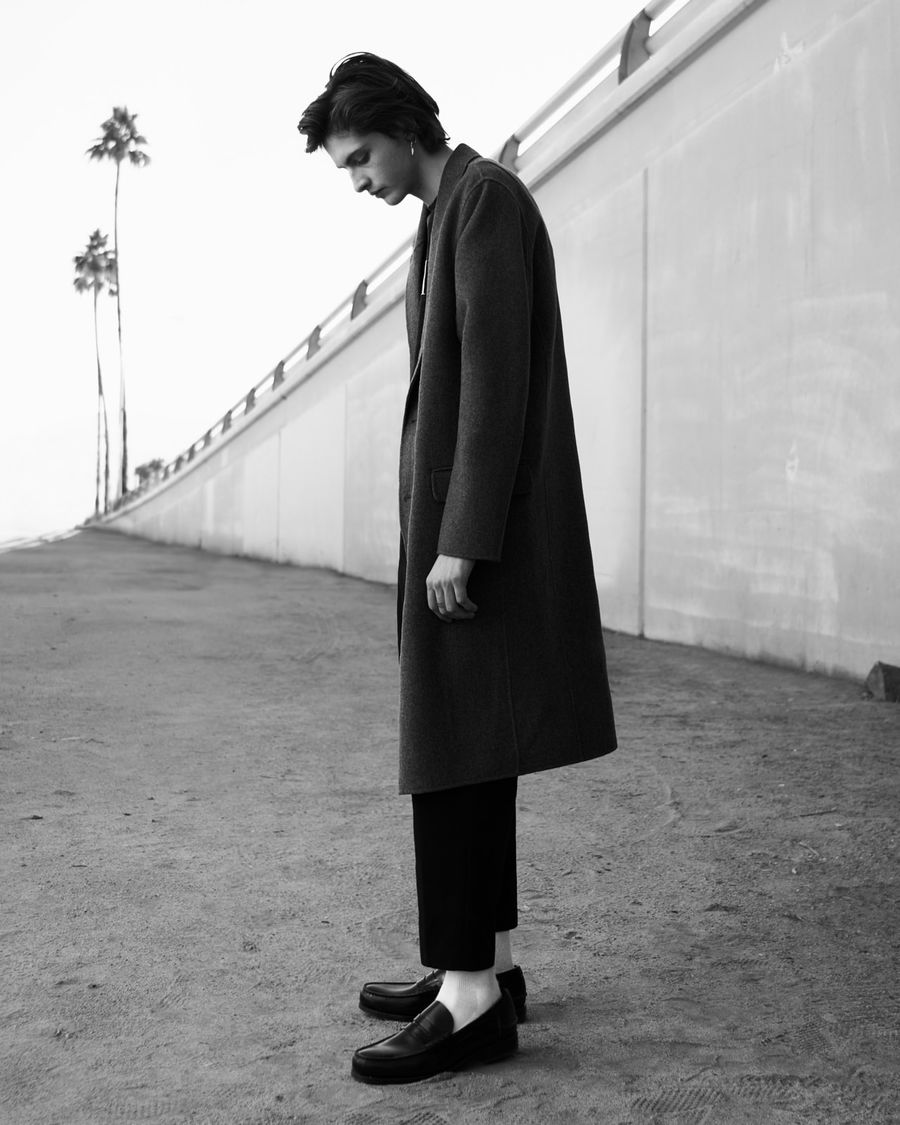 Black and white image of a man wearing a long grey coat with black tailored trousers and black loafers.