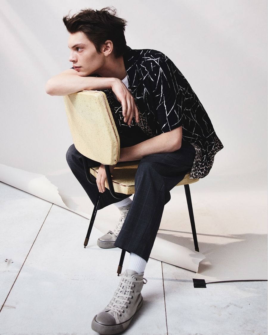 Immagine di un uomo seduto su una sedia con una camicia a fantasia. una t-shirt bianca, un paio di pantaloni scuri classici con trama a scacchi e un paio di sneakers grigie.