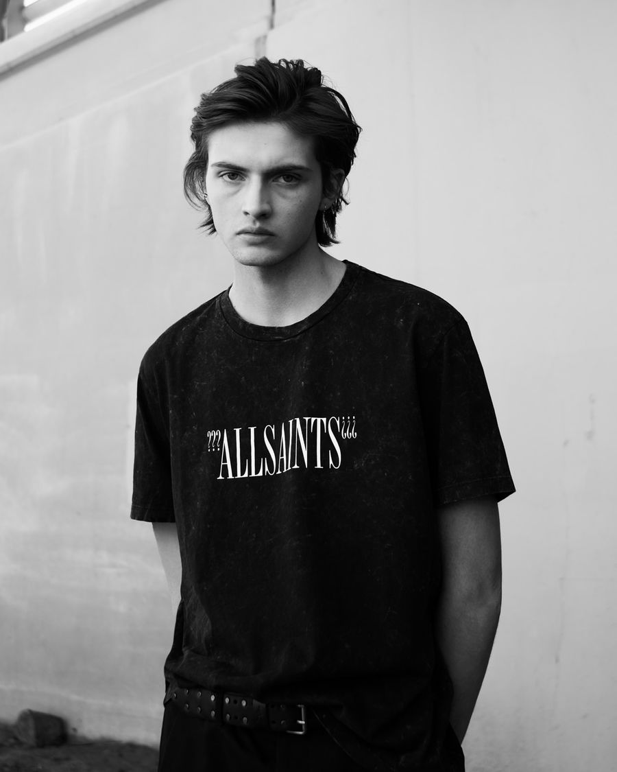 Imagen en blanco y negro de un hombre con una playera negra con un logo en blanco en el pecho y pantalones negros.