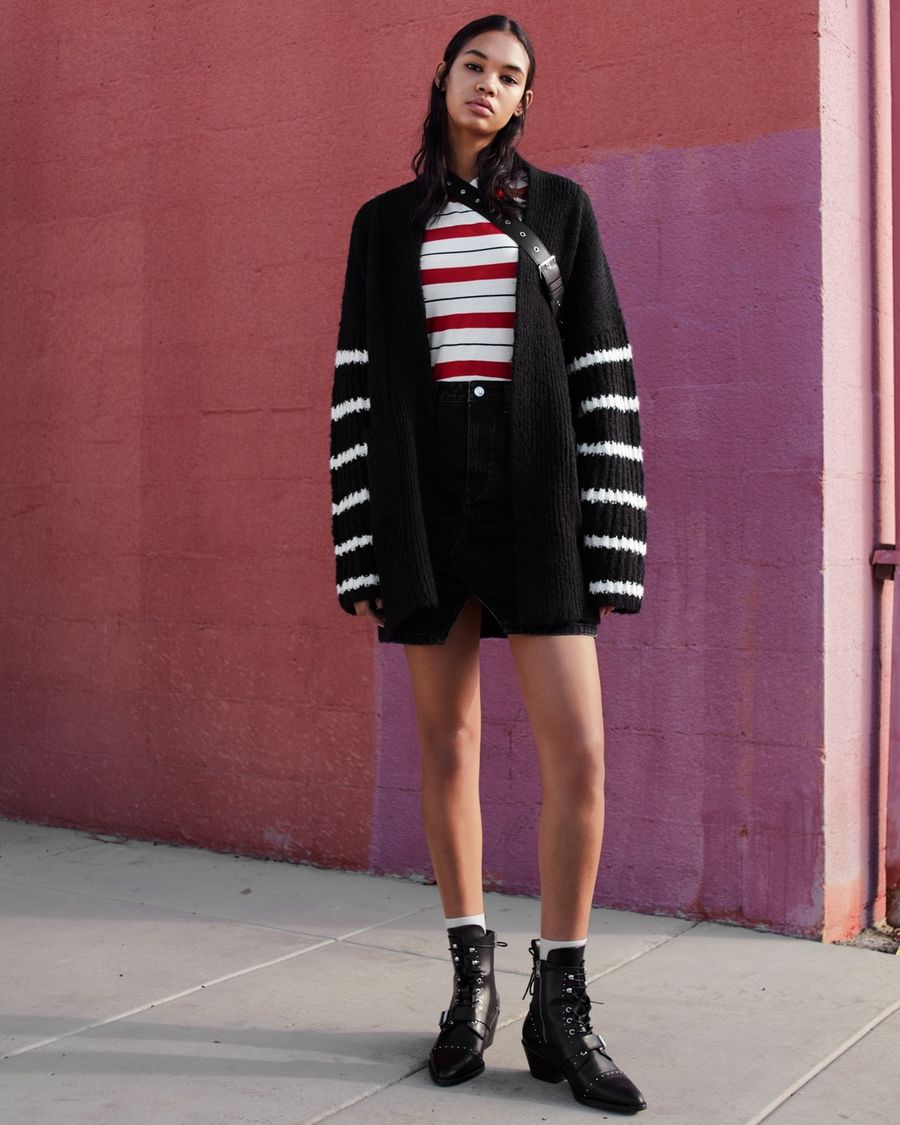 Woman standing in front of a pink wall wearing a short black denim skirt, white and red striped t-shirt, black cardigan white striped sleeves and black studded boots.
