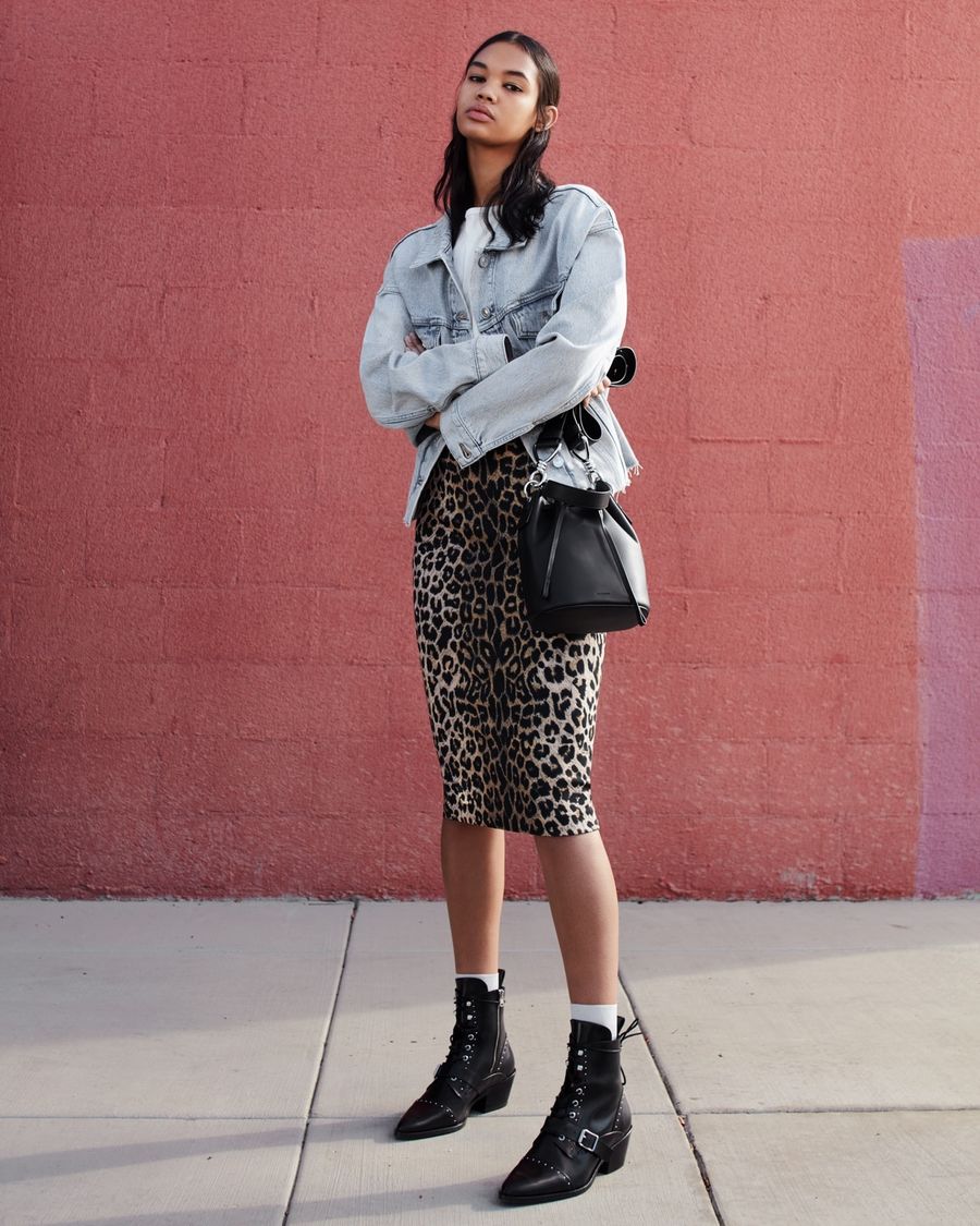 Image of a woman standing in front of a pink wall wearing a light denim shirt with a midi leopard print skirt, black leather boots and a black leather bucket bag.