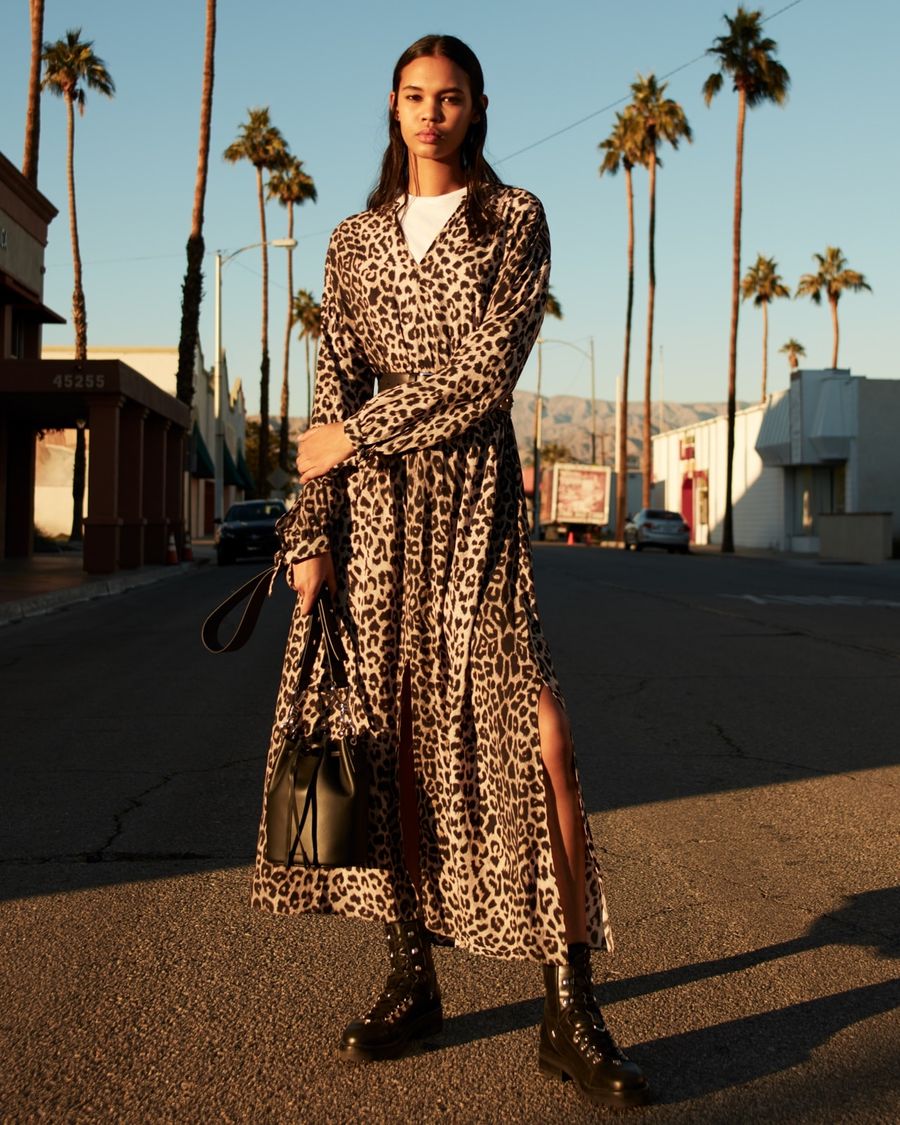 Image of a woman standing on a road wearing a long leopard printed dress over a white t-shirt, with black leather boots and a black leather bucket bag.