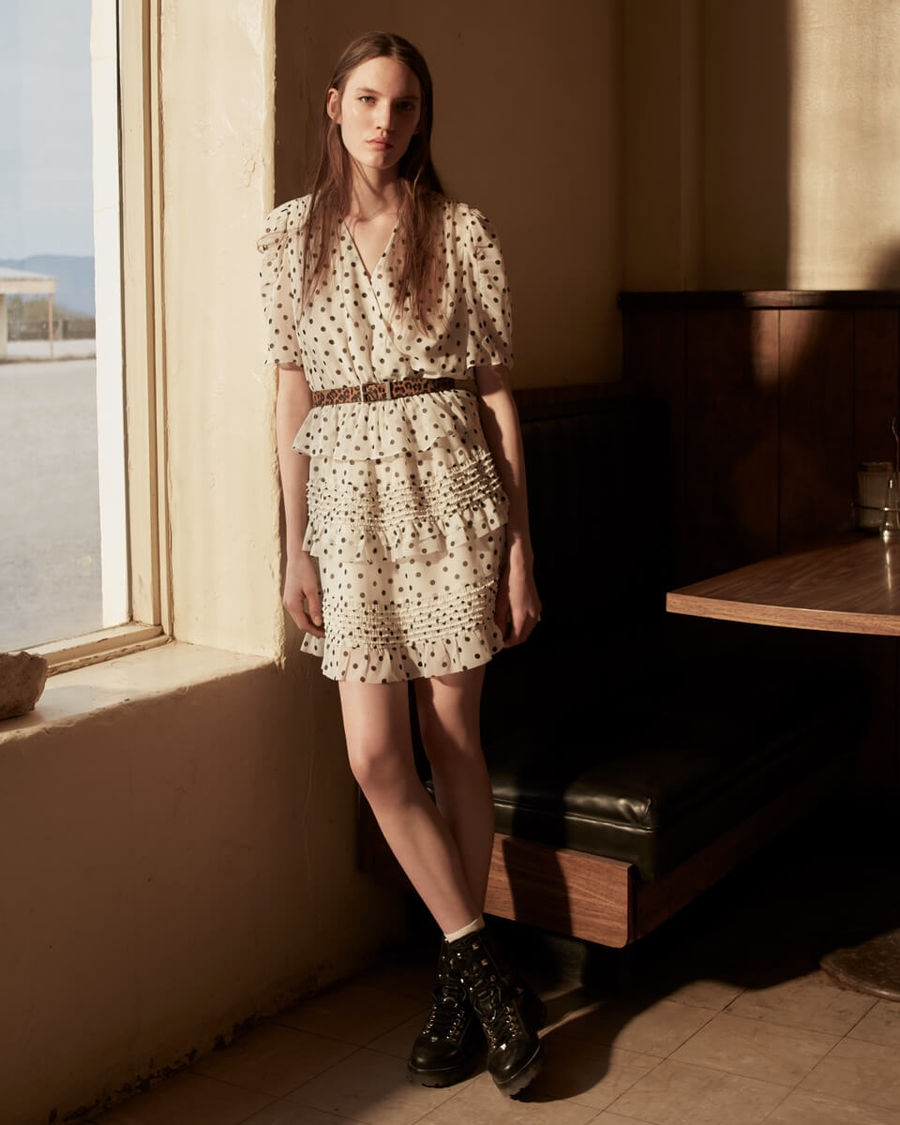 Woman resting against a wall near a window and wearing a dot printed beige dress with a leopard print belt and black leather boots.