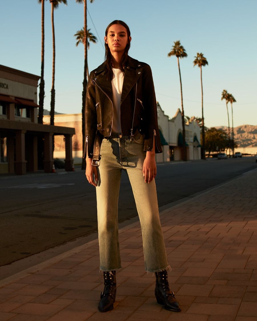 Placeholder image of a woman standing in the street at sunset wearing a black leather jacket with a white t-shirt, light blue denim jeans and black leather boots.