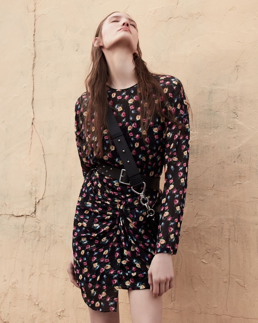 Woman standing in front of a faded yellow wall wearing a flower printed long sleeve dress with black leather studded belt and crossbody bag.