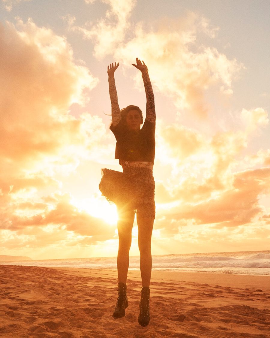 Image of a woman jumping on the beach at sunset wearing a short sleeve plain t-shirt over a printed long t-shirt, a printed skirt and black leather boots.