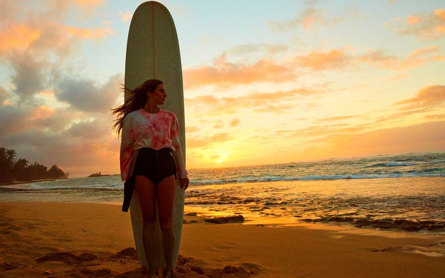 Image of a woman standing on the beach at sunset in front of a surf board and wearing a long sleeve pink tie dye top with her wet suit.