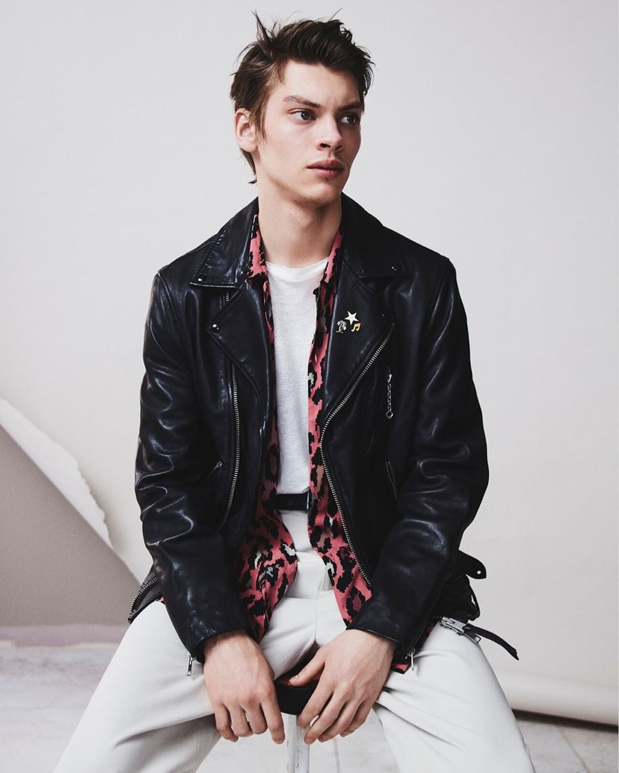Portrait of a man wearing a black leather jacket, pink leopard printed shirt, white t-shirt and white jeans.