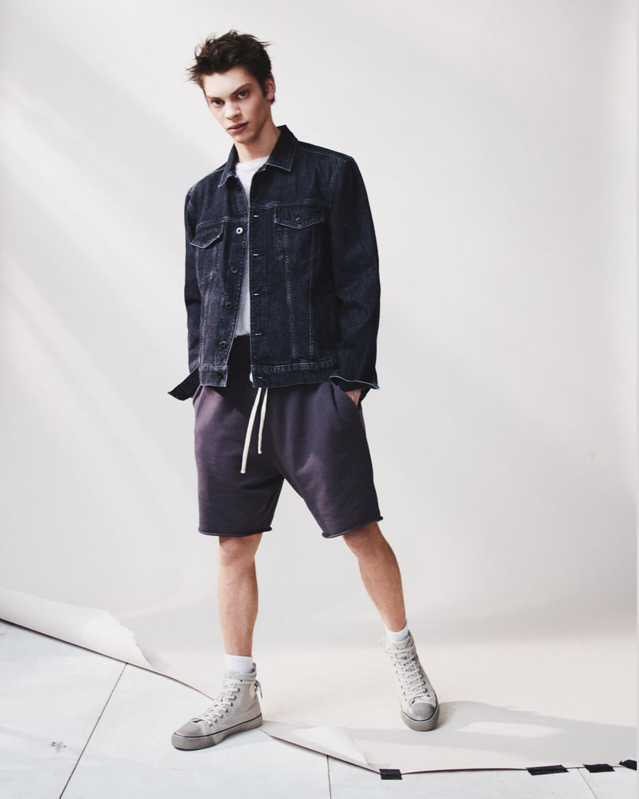 Image of a man wearing a dark denim jacket with a white t-shirt, dark sweatshorts and white sneakers.