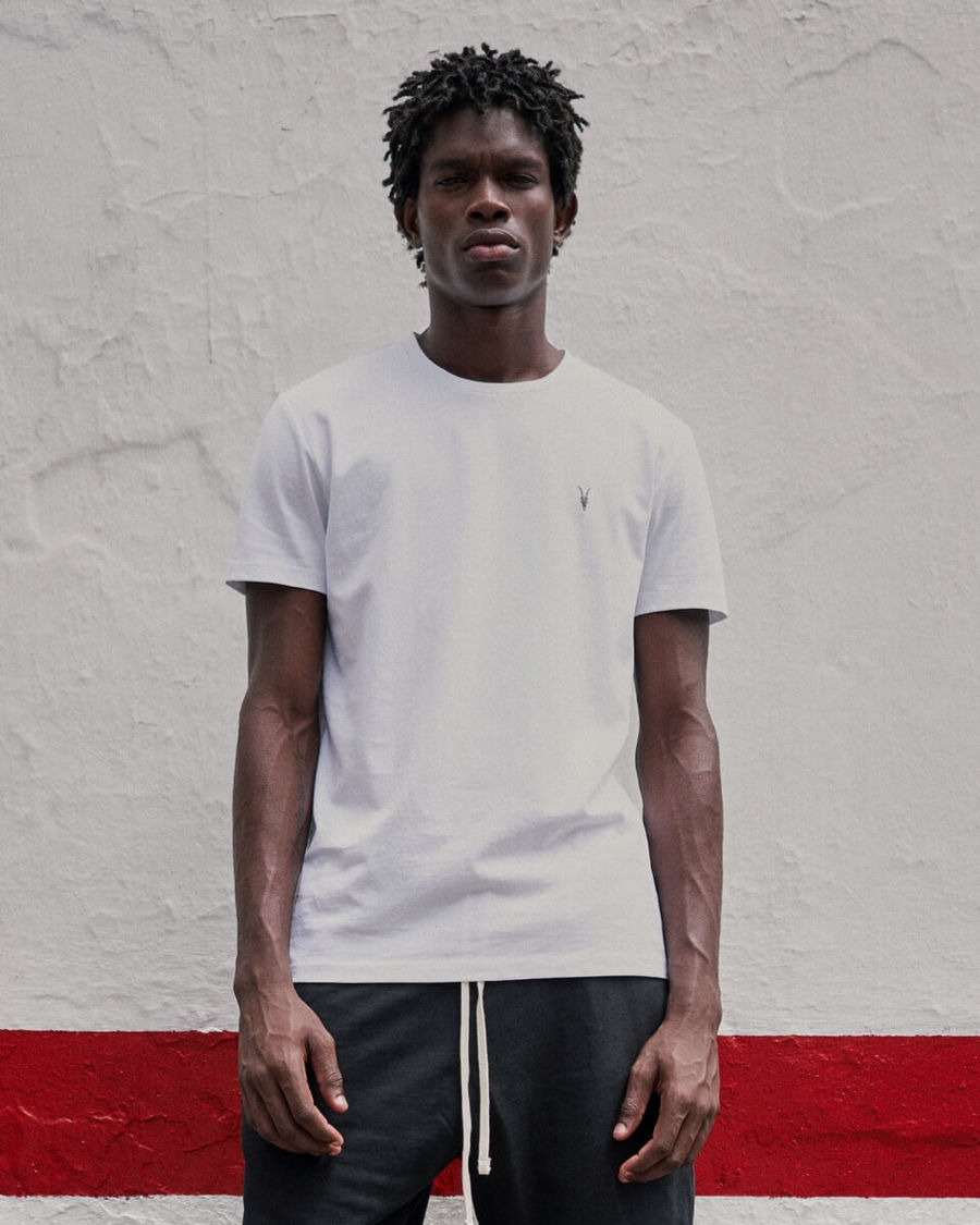 Portrait of a man standing in front of a white and red wall wearing a white t-shirt from our Ramskull Collection with dark sweatpants.