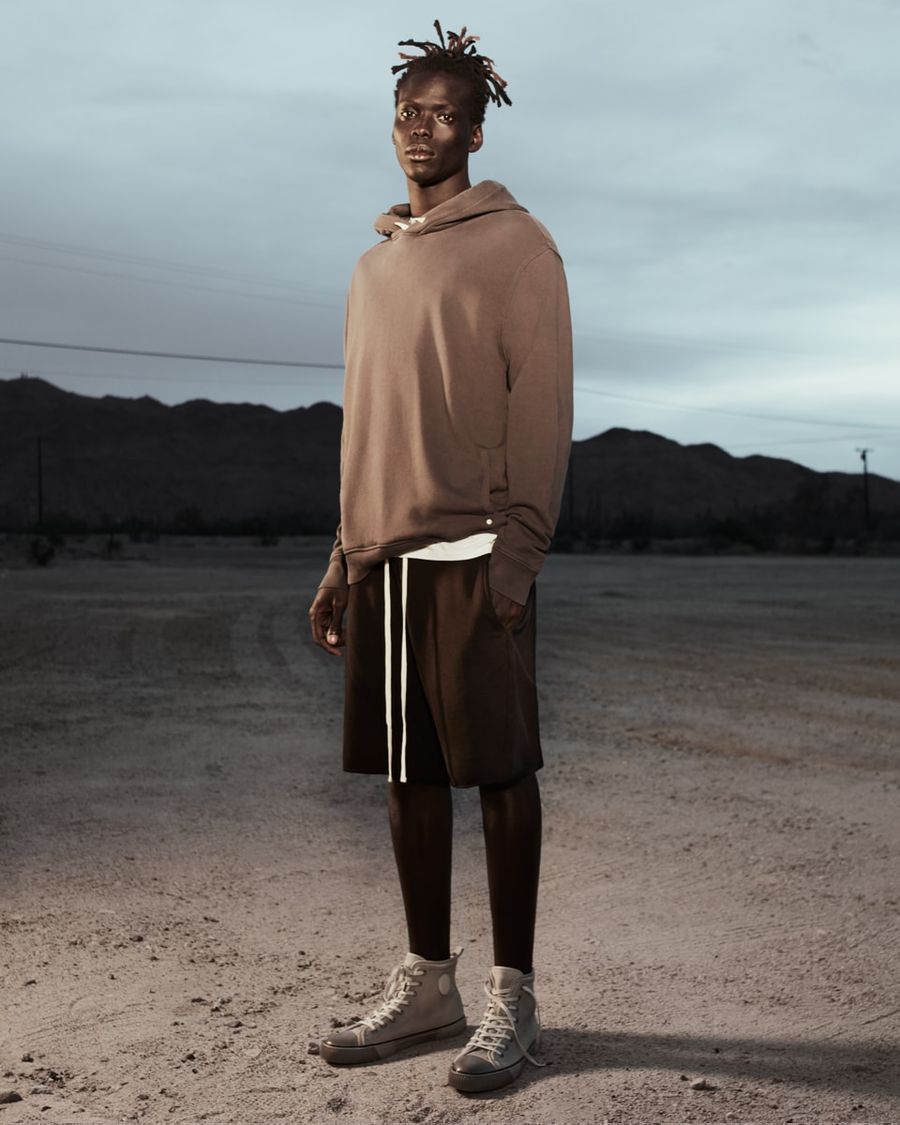 Image of a man standing in the desert at sunset wearing a sand coloured hoodie with black shorts and white trainers.