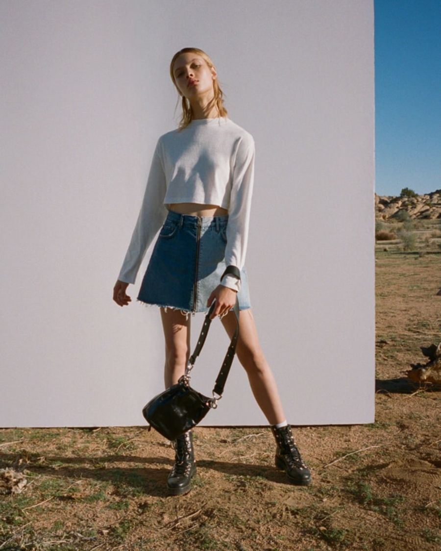 Image of a woman wearing a white cropped long sleeve t-shirt with a denim skirt, black leather boots and holding a black leather bag.